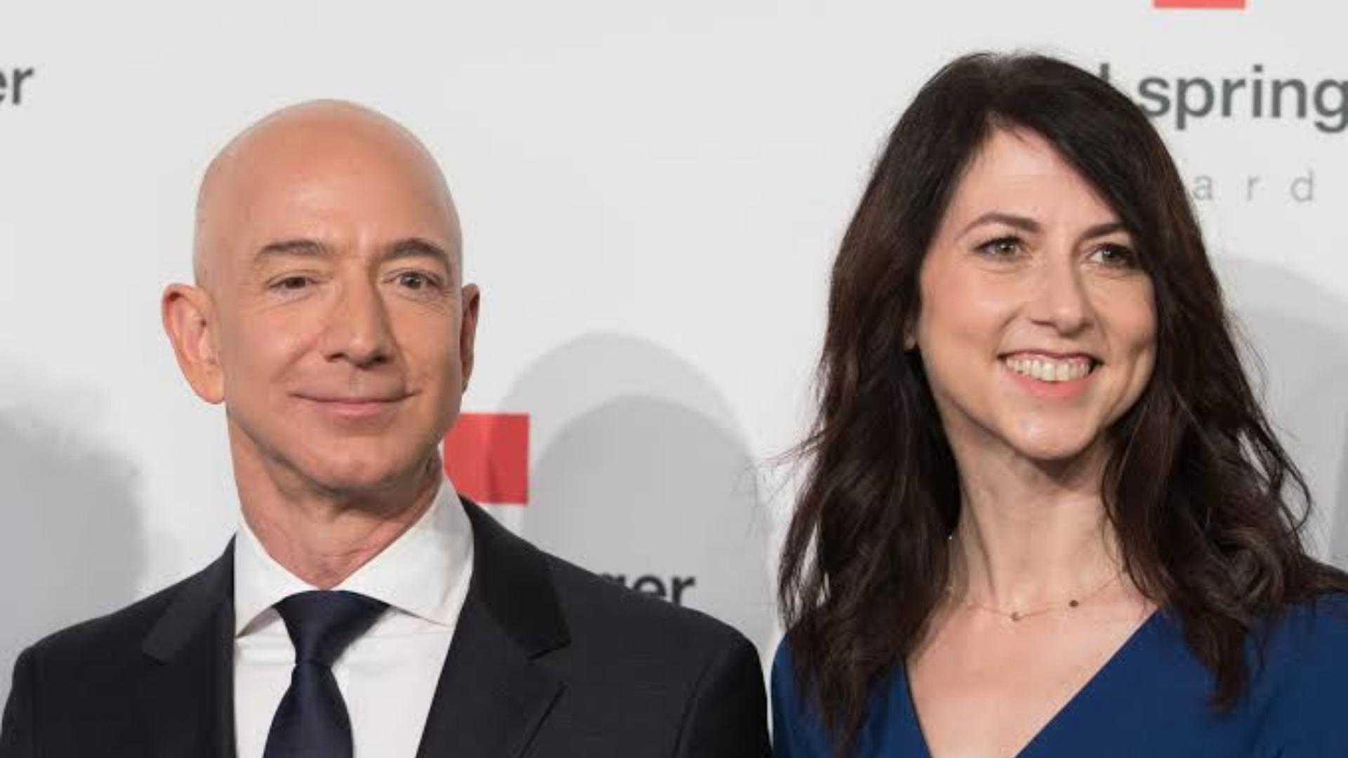 jeff bezos in a dark suit and tie standing beside a smiling mackenzie scott in a blue dress at a formal event backdrop
