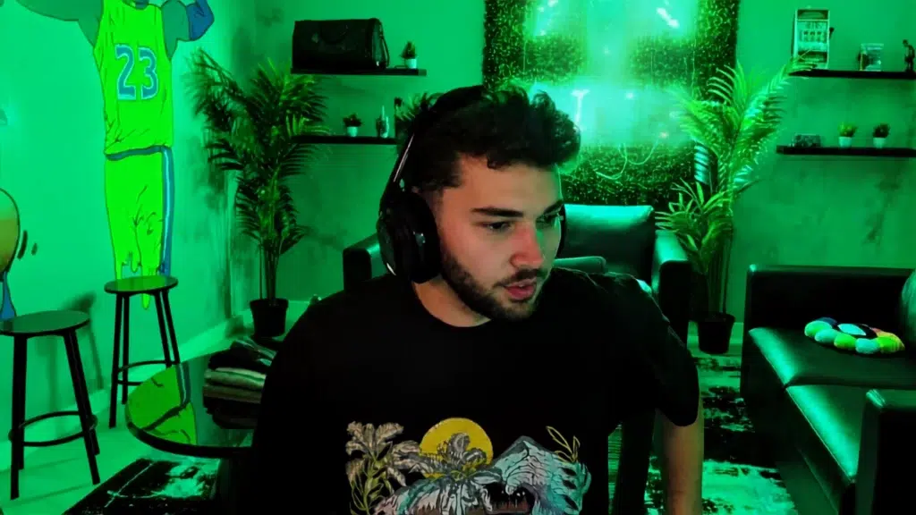 young male streamer wearing headphones and a black graphic t-shirt in a green-lit streaming room with plants and lounge furniture