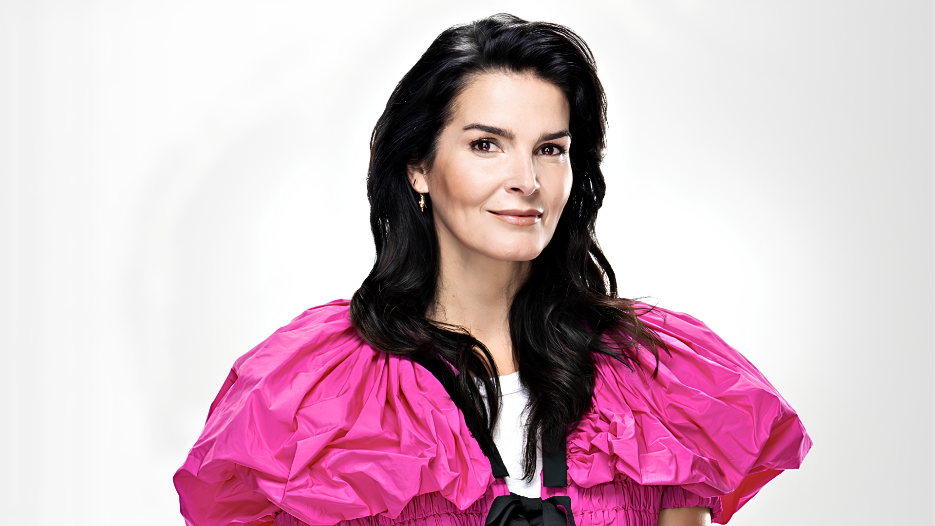 Angie Harmon with dark, wavy hair posing in a bright pink puffed-sleeve top against a white backdrop.