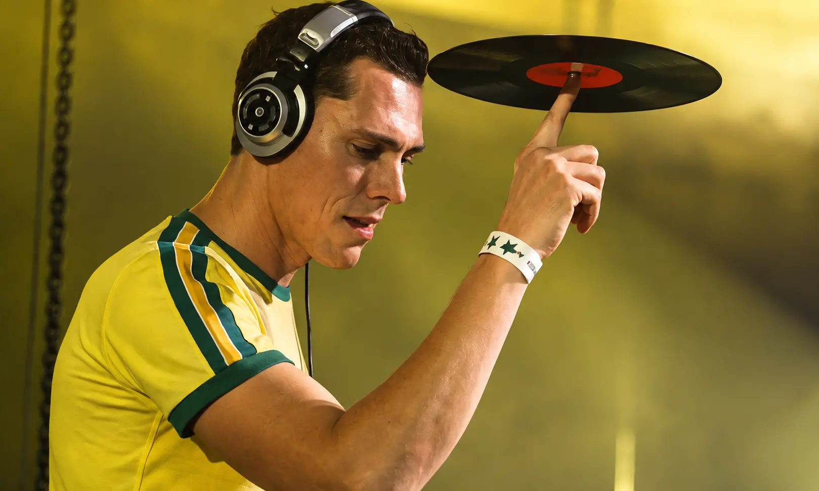 DJ Tiësto wearing headphones while balancing a vinyl record during a live performance with warm stage lighting and club atmosphere.