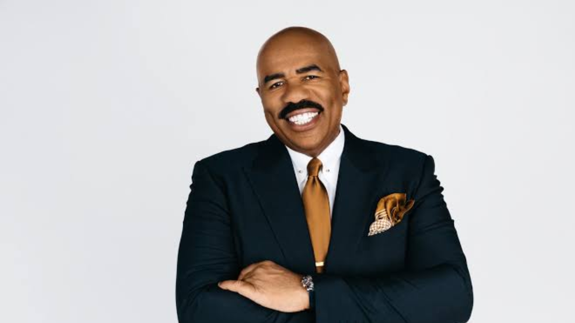 Steve Harvey smiling in a black suit, white shirt, and gold tie with arms crossed.