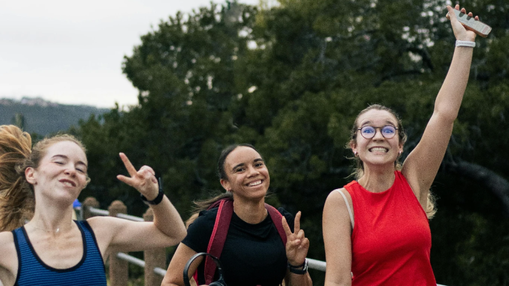 Three women jogging and smiling on a park pathway surrounded by greenery