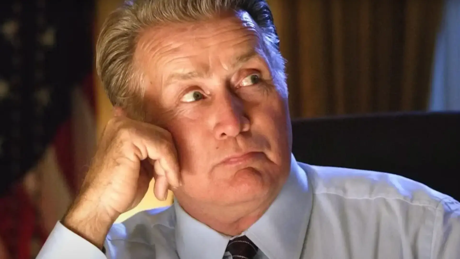 Martin Sheen in a light blue shirt and dark tie resting his head on one hand while looking upward indoors.