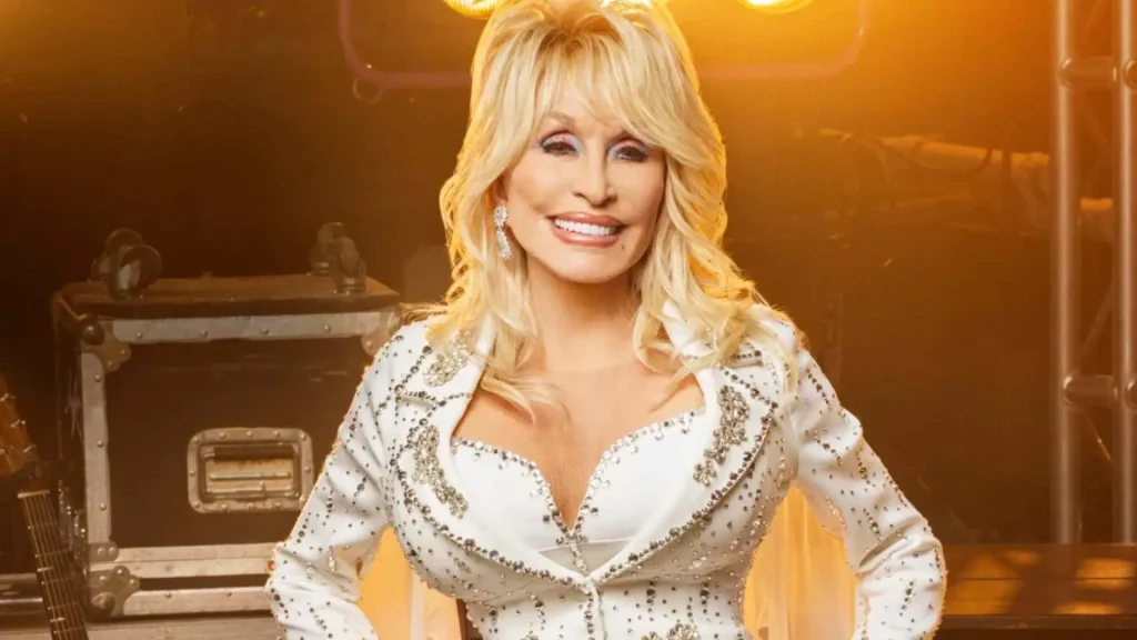 dolly parton smiling on stage in a white embellished outfit with blonde hair and sparkling earrings under warm lights