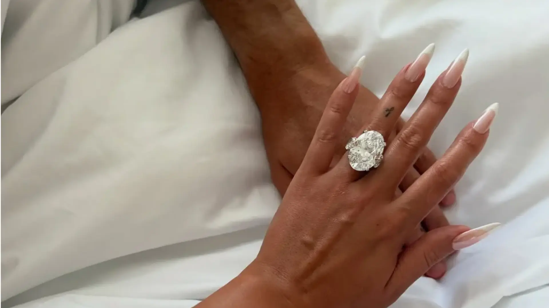 close-up of a manicured hand wearing a large oval diamond engagement ring with side stones against soft white fabric