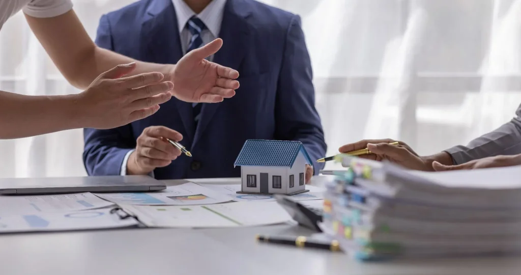 Business meeting with model house on table surrounded by paperwork and hands gesturing in discussion