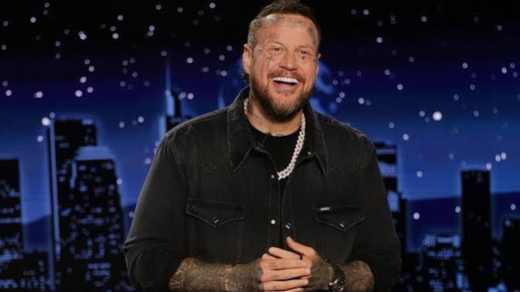 smiling jelly roll in black jacket and chain necklace seated before a blue nighttime city skyline with scattered lights