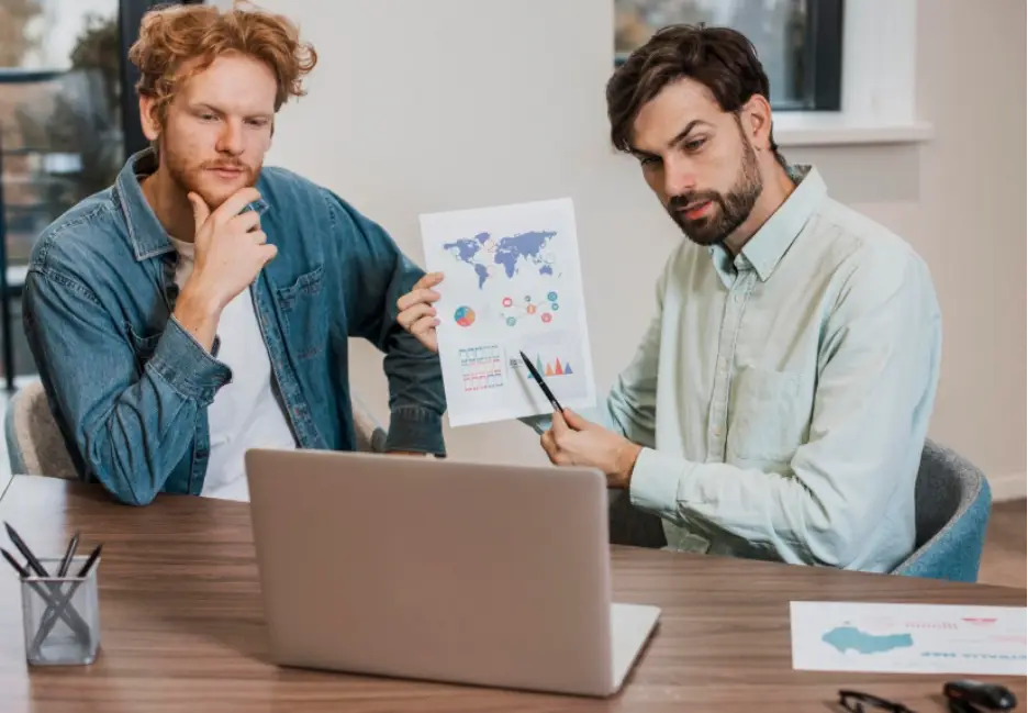 Two men discussing charts and graphs on laptop in modern office setting