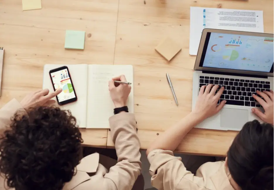 Two people reviewing analytics on laptop and smartphone at wooden desk with notebook