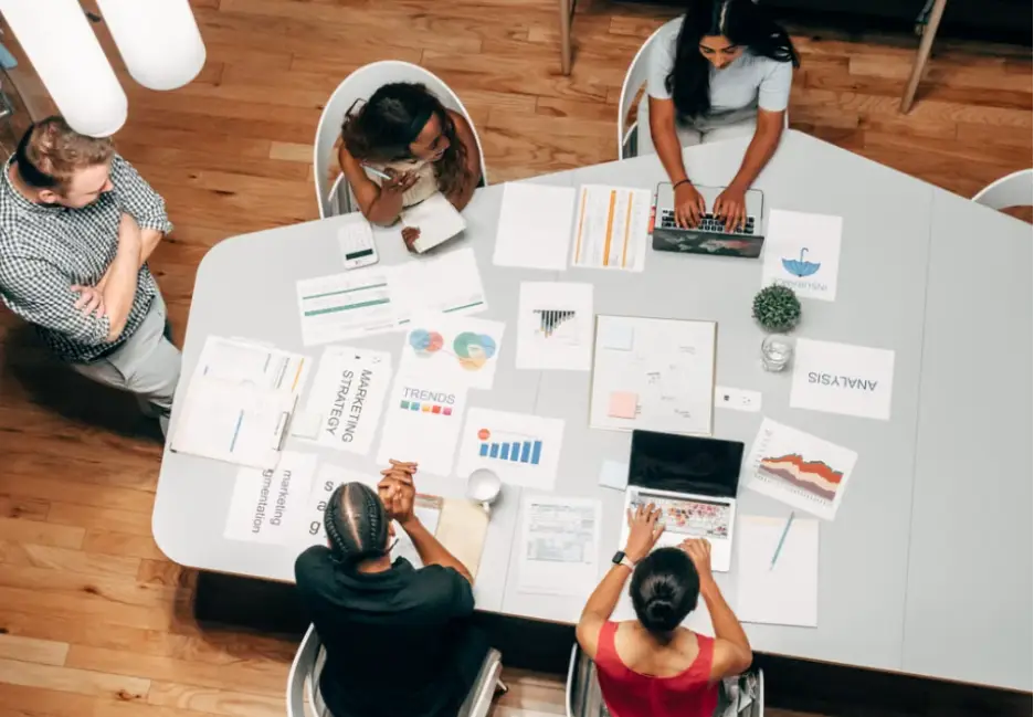 Team collaborating around a table with laptops and marketing strategy documents in an office setting