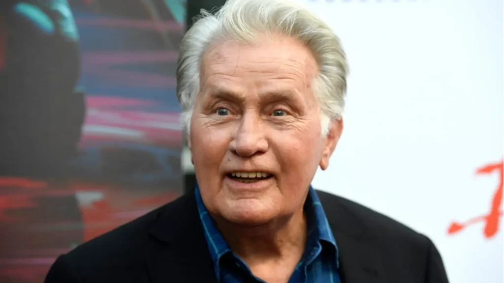 martin sheen smiling at a red carpet event in a black blazer and blue shirt against a bright event backdrop