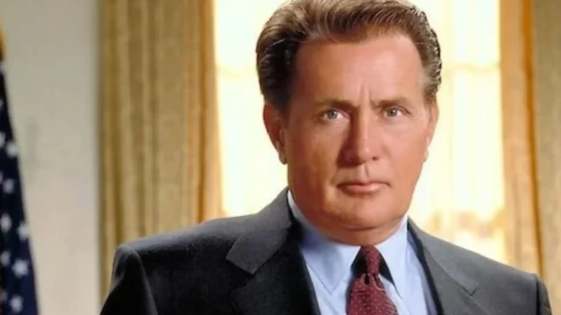 Martin Sheen in a dark suit, light blue shirt, and red tie seated indoors with bright curtains and a serious expression