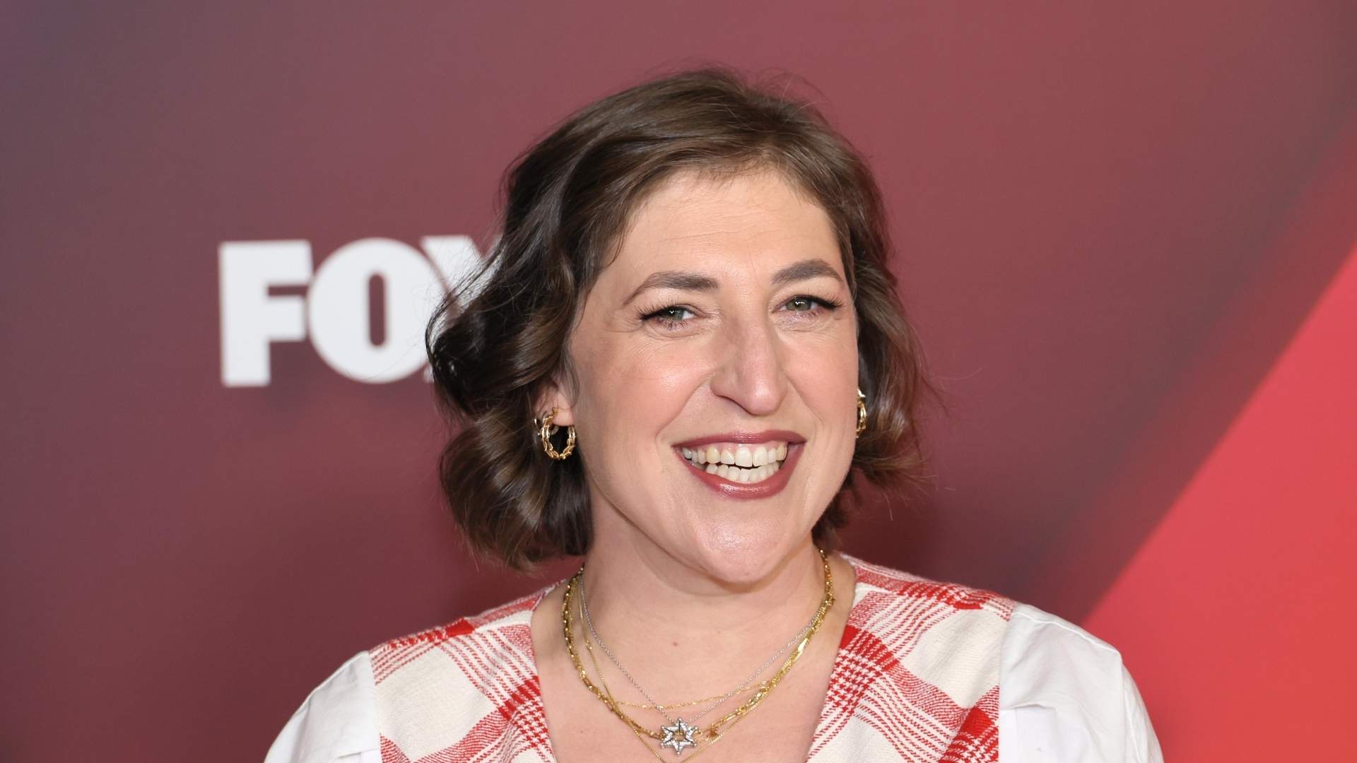 Mayim Bialik smiling on a red carpet wearing a red and white plaid vest with gold necklaces.