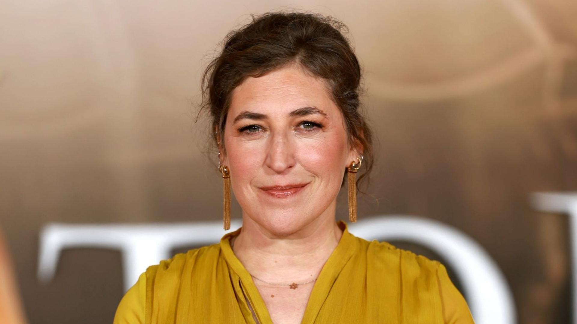 Mayim Bialik wearing a mustard yellow dress and gold earrings at an indoor event.