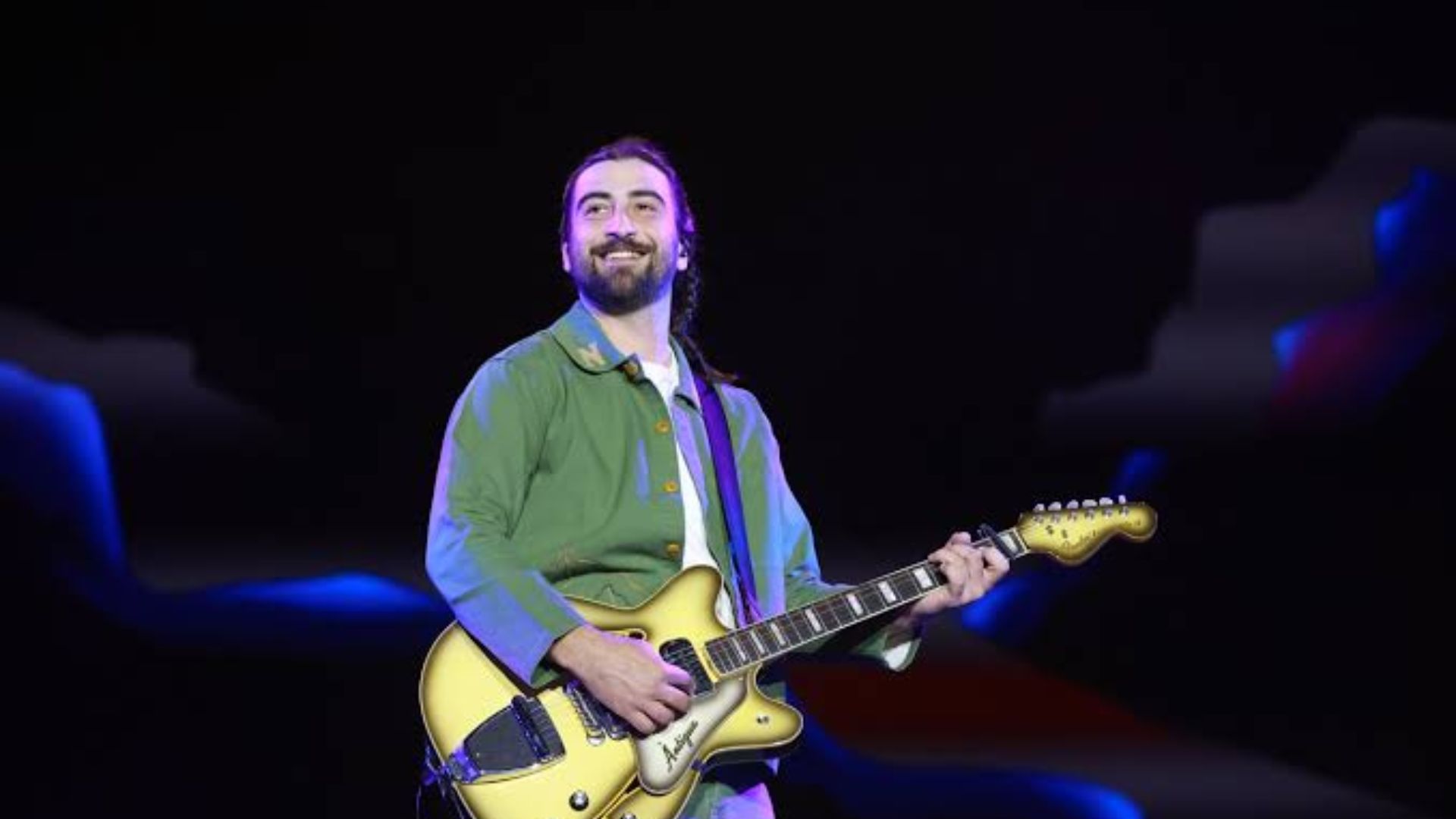 Noah Kahan smiling on stage while playing an electric guitar during a live performance.