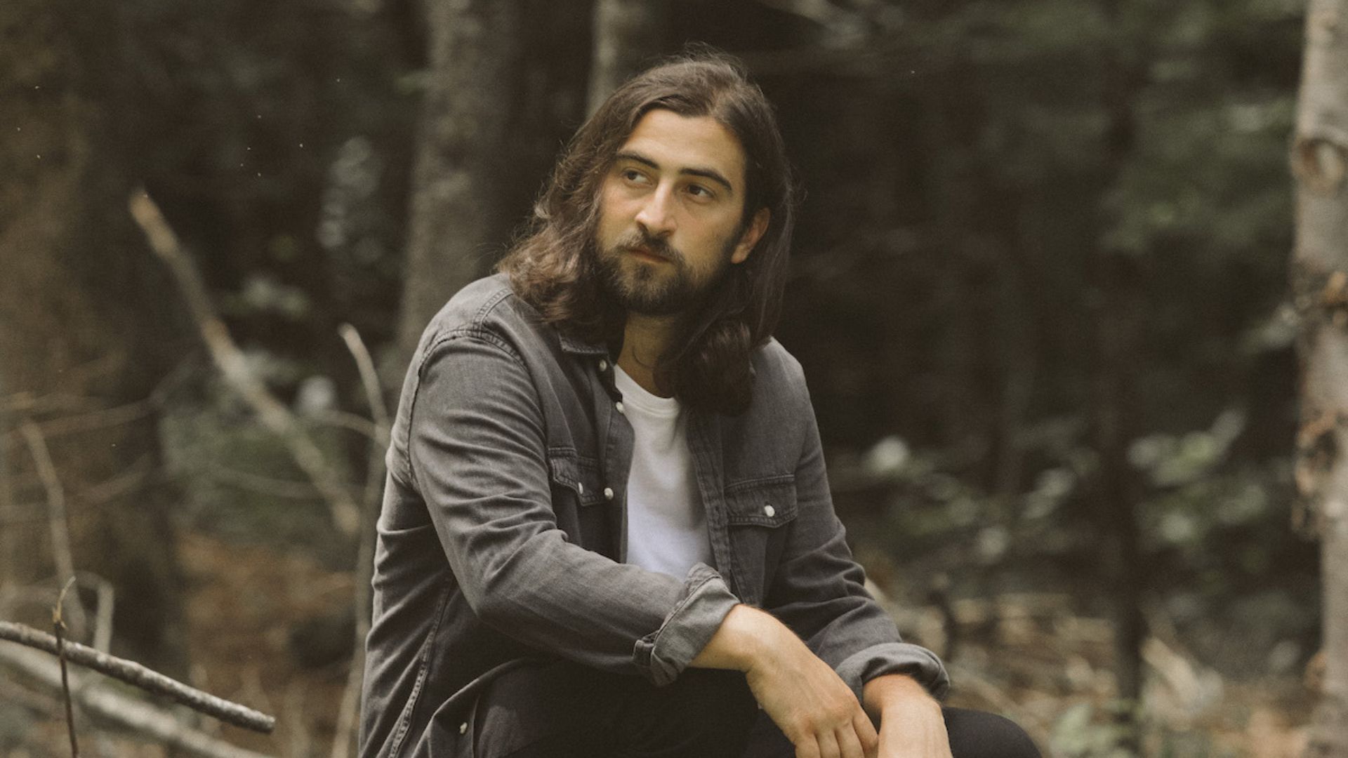 Noah Kahan sitting outdoors in a wooded area wearing a dark jacket over a white shirt.