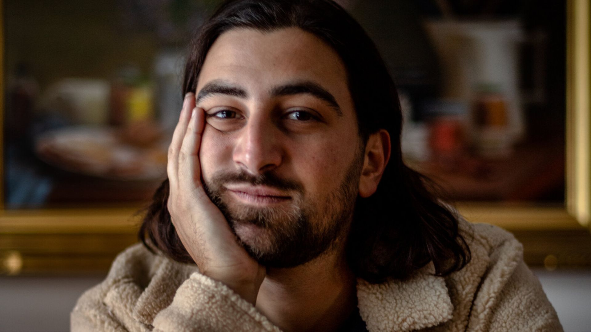 Noah Kahan resting his face on his hand while sitting indoors in a casual portrait.