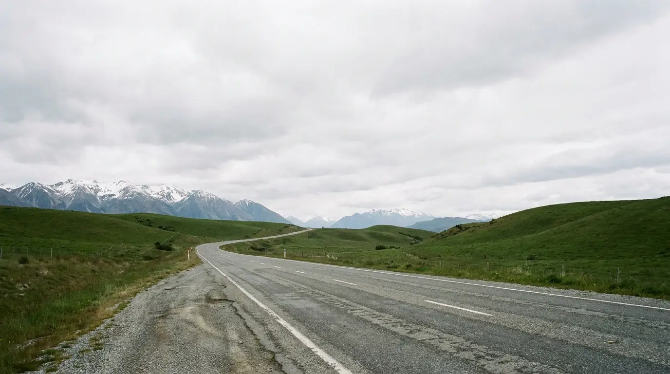 Winding road stretching through lush green hills with snow-capped mountains under overcast sky