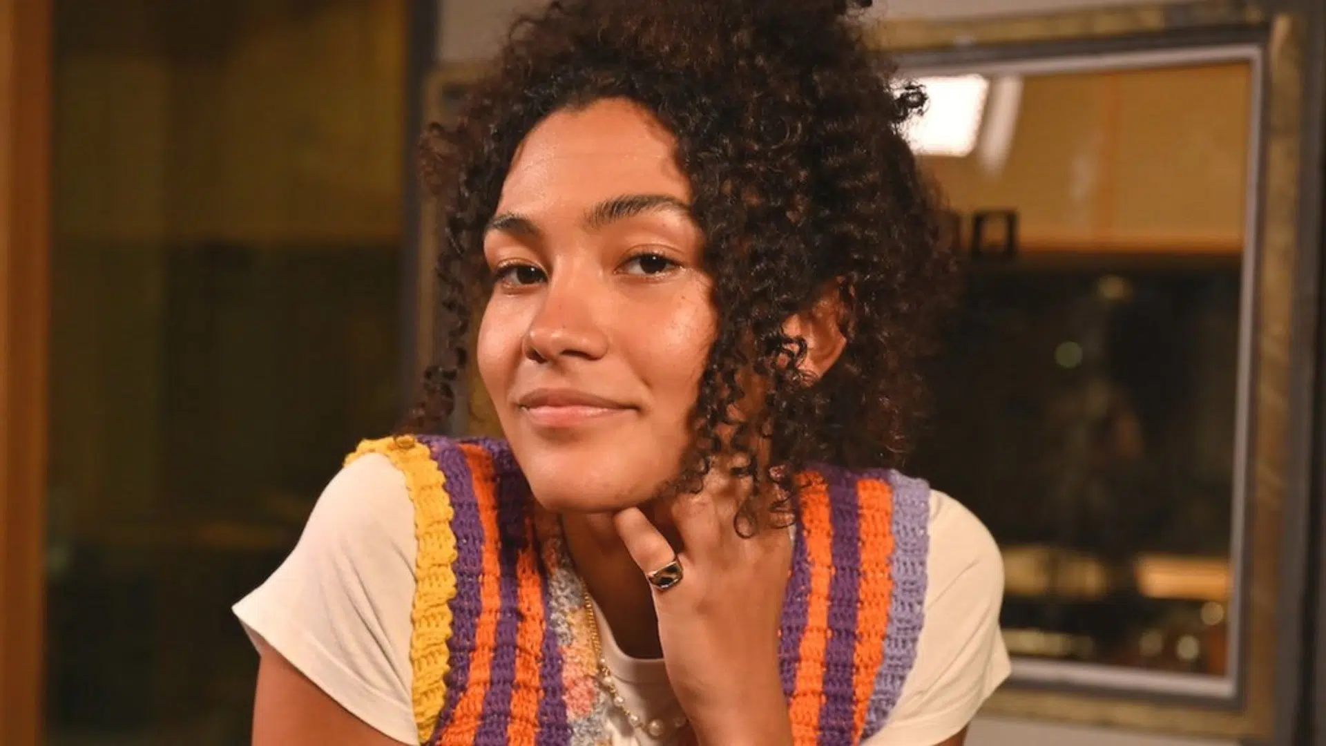 olivia dean smiling in a close up portrait wearing a colorful knitted vest indoors with warm lighting.