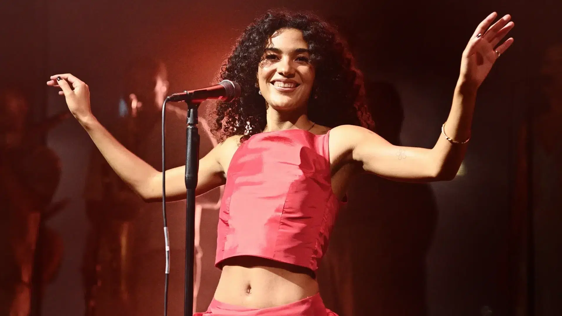 olivia dean performing live on stage wearing a pink outfit with microphone and warm concert lighting.