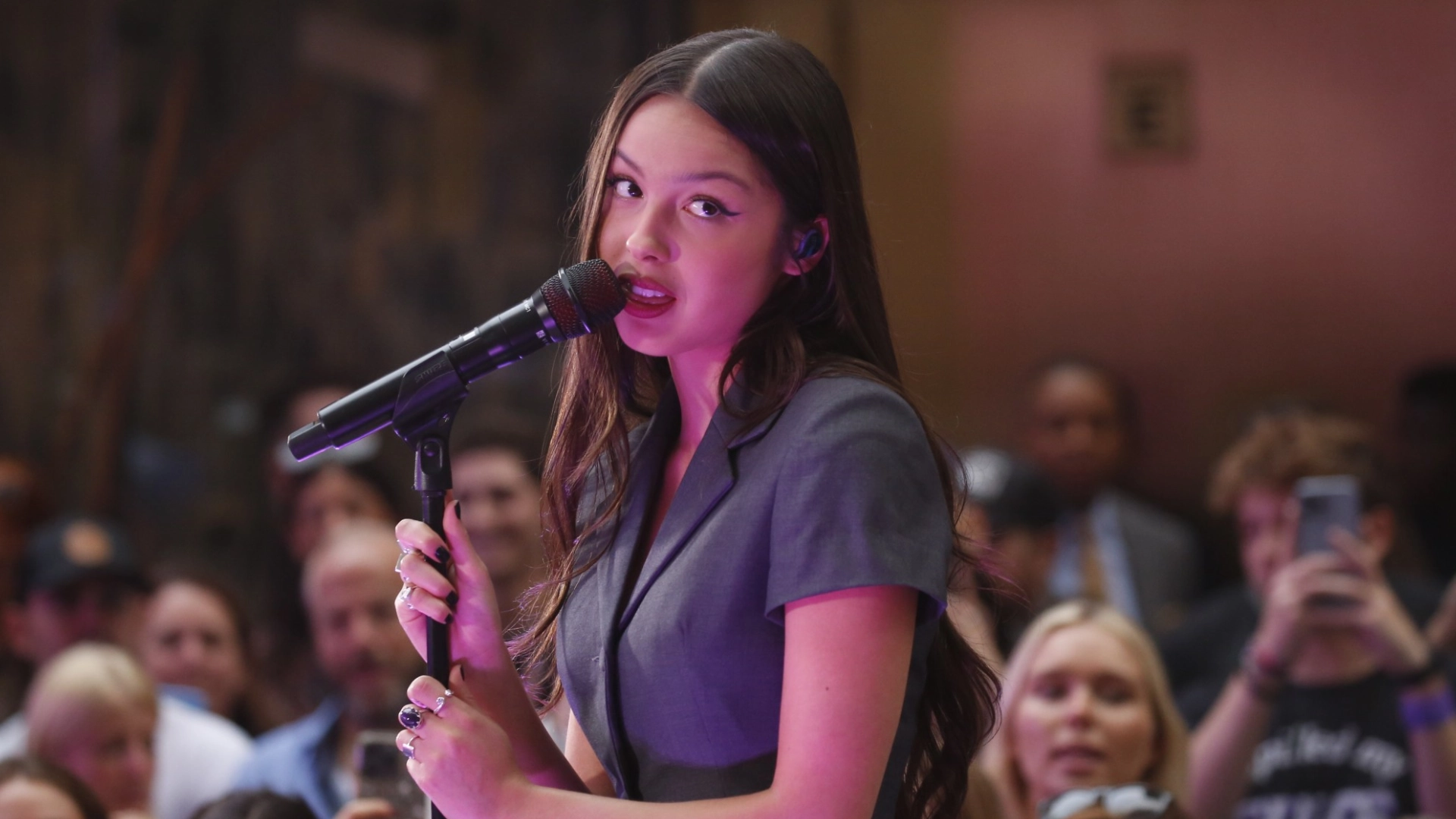 olivia rodrigo performing on stage with a microphone, audience in background, and purple concert lighting.