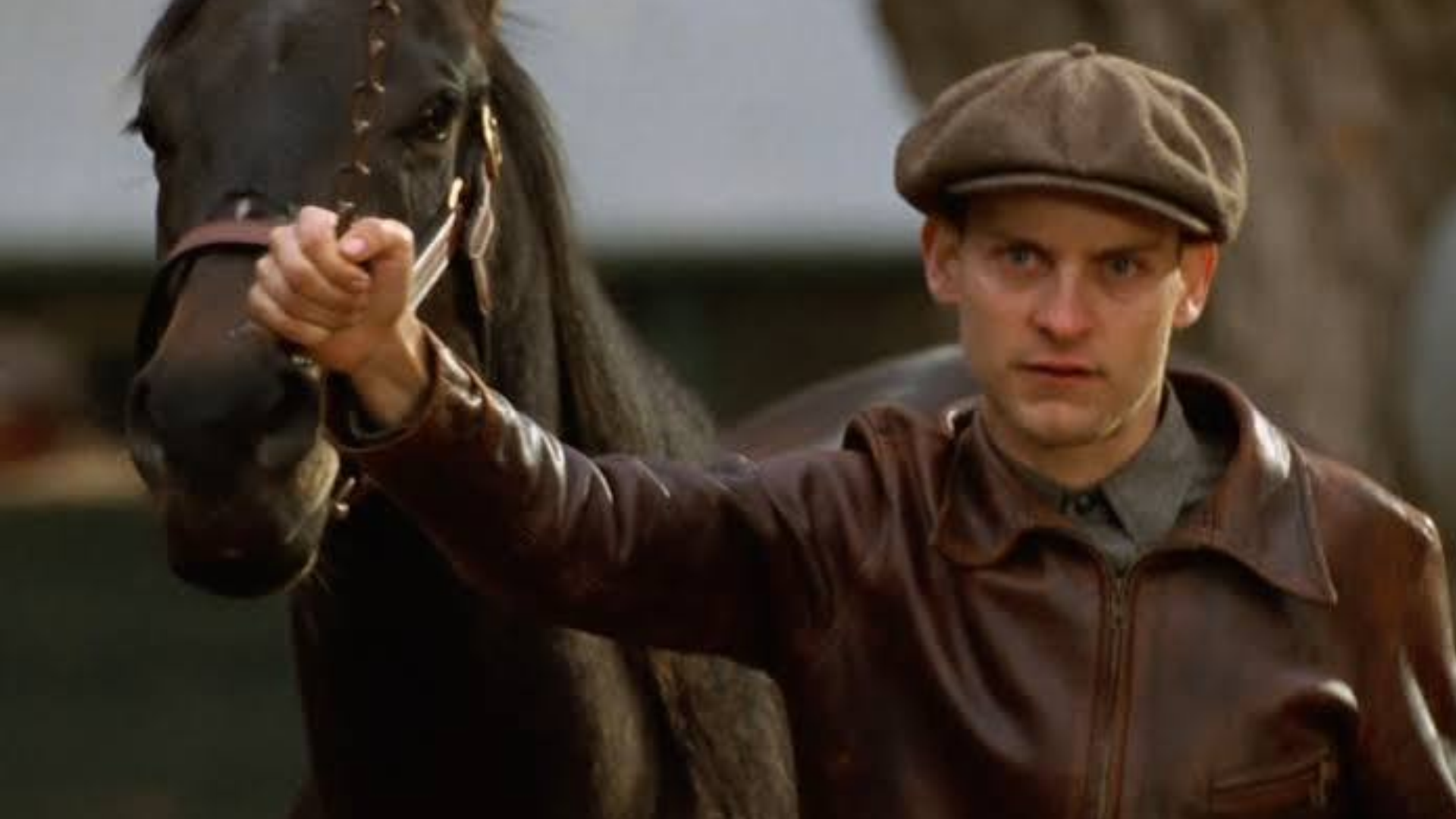 tobey maguire in brown leather jacket and flat cap holding the reins of a dark horse while standing outdoors beside a stable building