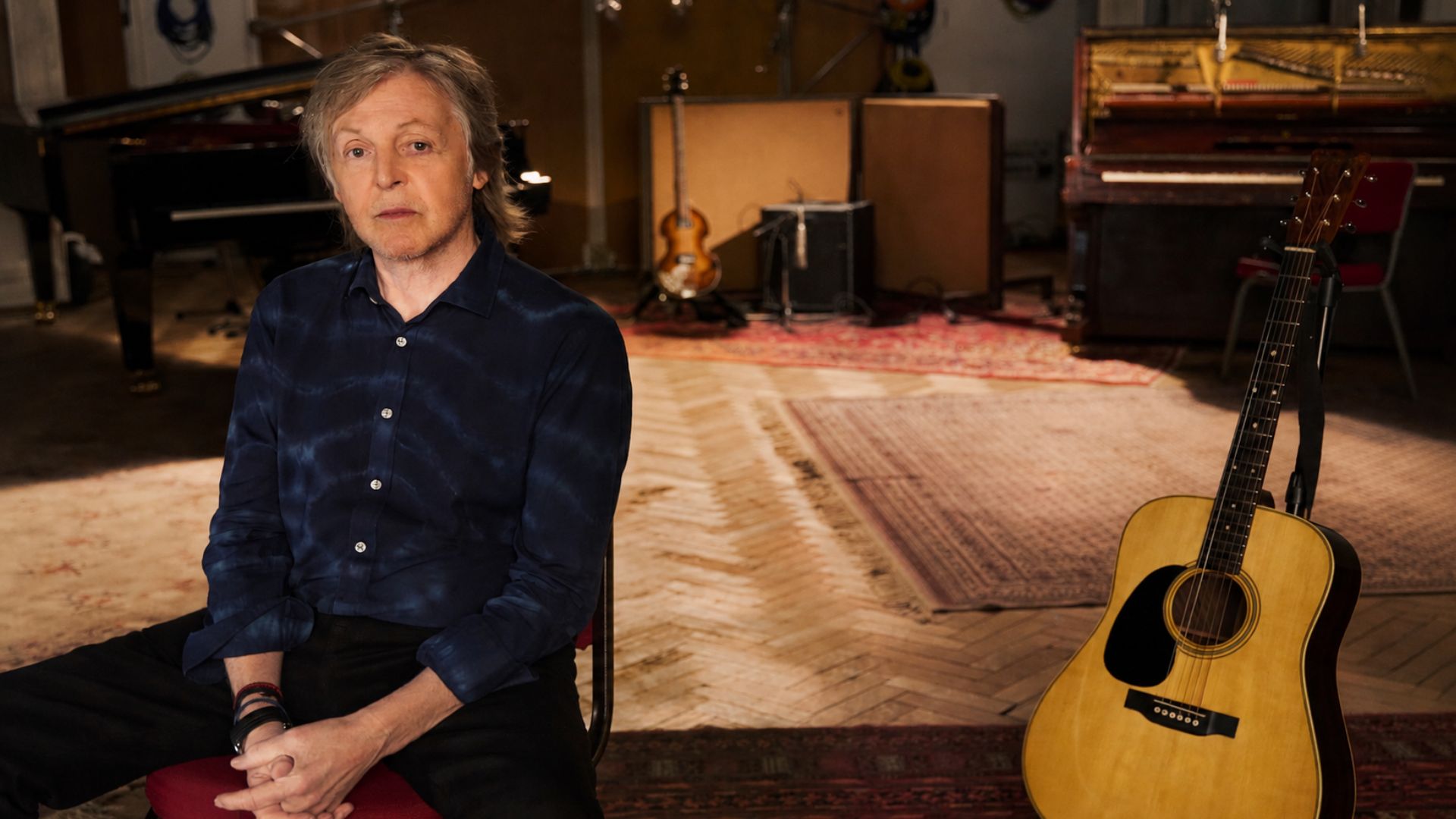 Paul McCartney sitting in a recording studio with an acoustic guitar nearby and musical instruments in the background.