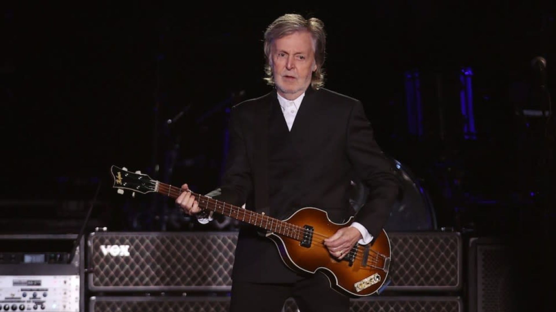 Paul McCartney performing on stage in a black suit while playing his signature bass guitar under concert lights.