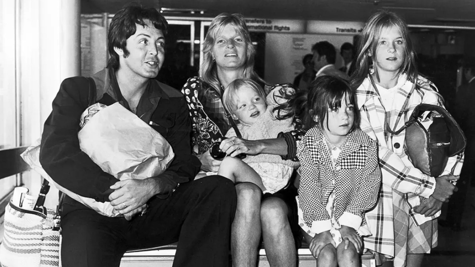 Black-and-white photo of Paul McCartney seated with his children and family at an airport.