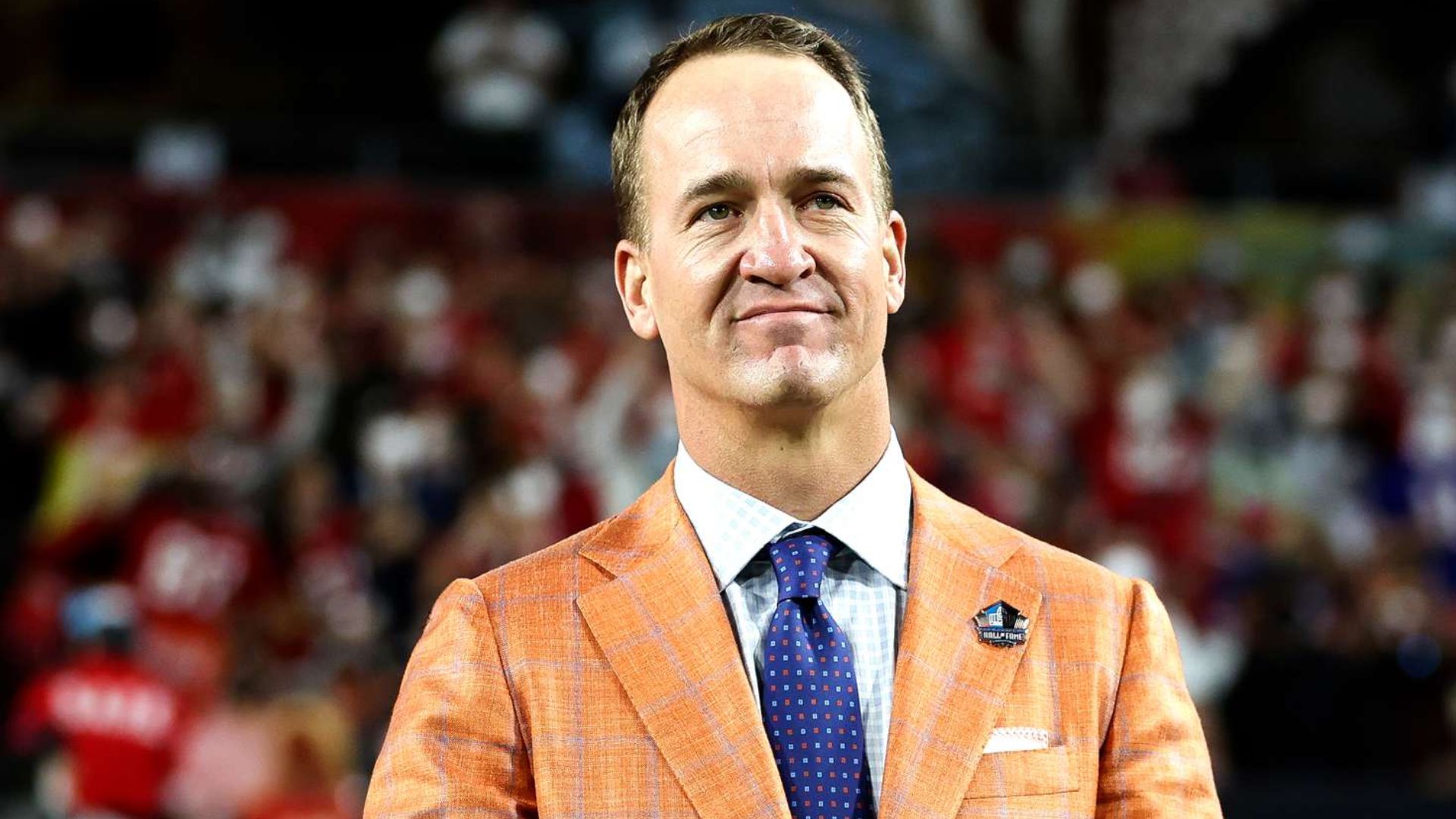 Peyton Manning wearing an orange blazer and tie while standing on a football field during an event.