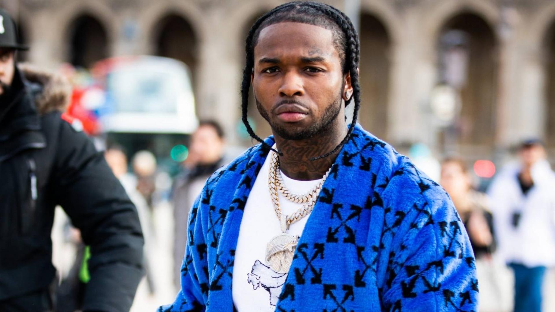 Pop Smoke, an American rapper, wearing a blue patterned jacket, gold chains, and standing outdoors in a city setting.