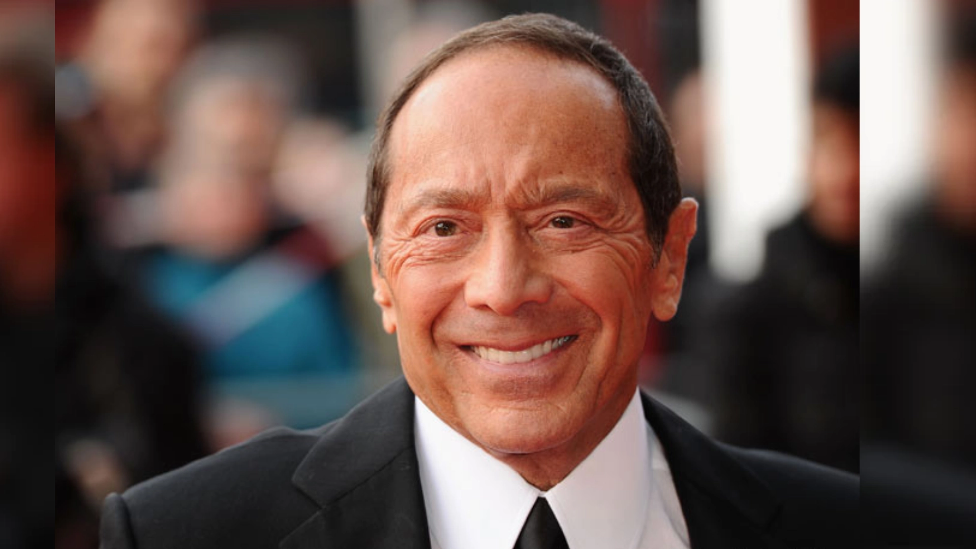 paul anka wearing a black suit and tie smiling at a public event with blurred background