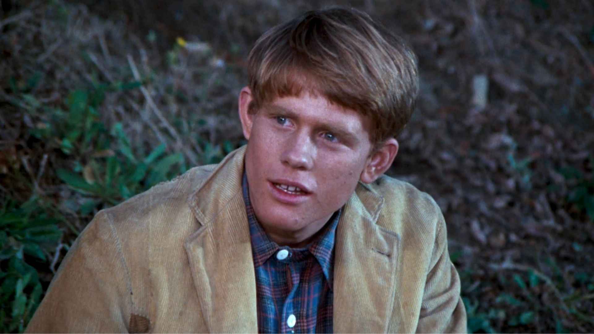 Young Ron Howard in a tan corduroy jacket and plaid shirt, seated outdoors in a scene from a classic film or TV show.