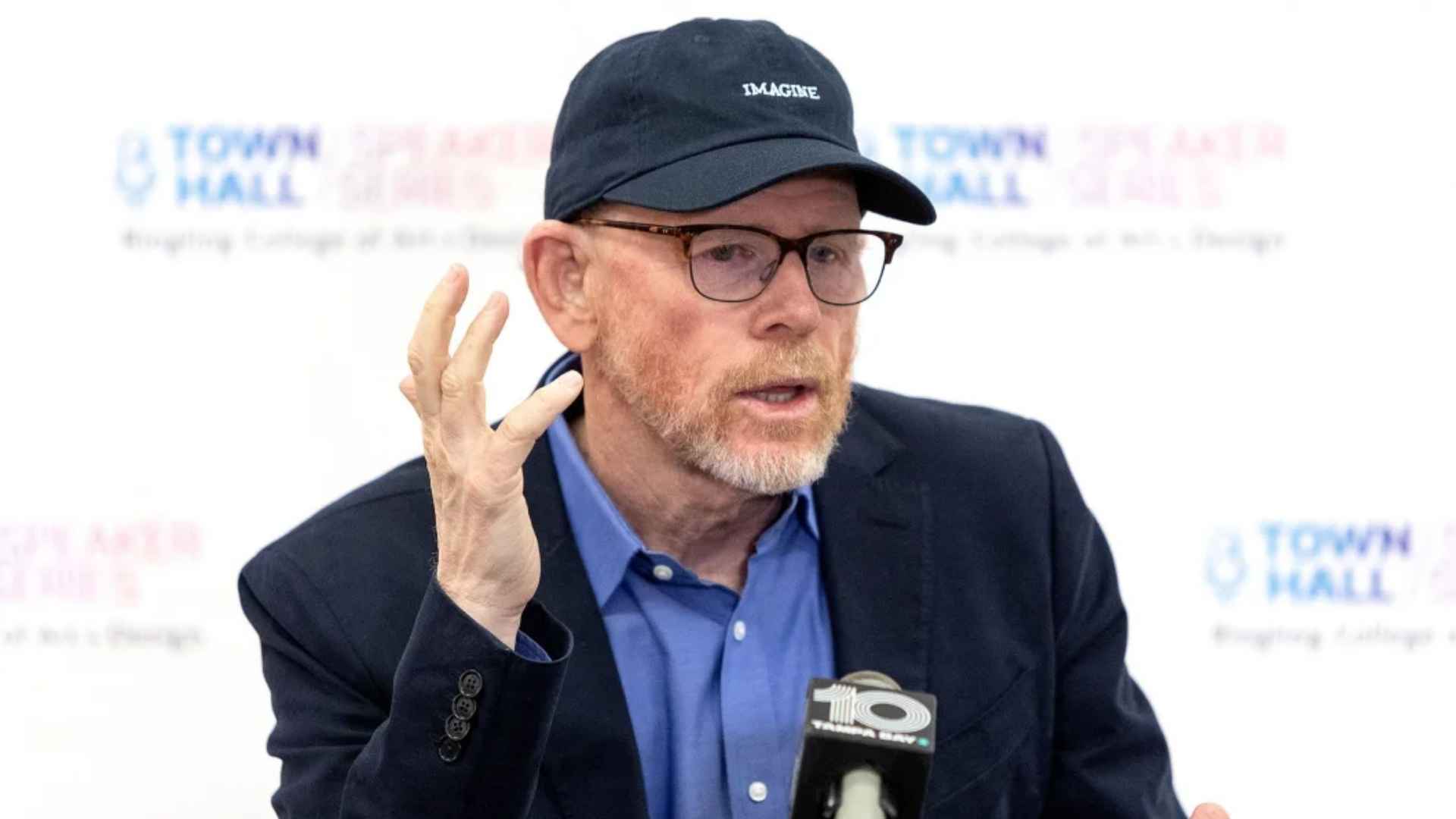 Ron wearing glasses and a navy cap speaking into a microphone during a press event.