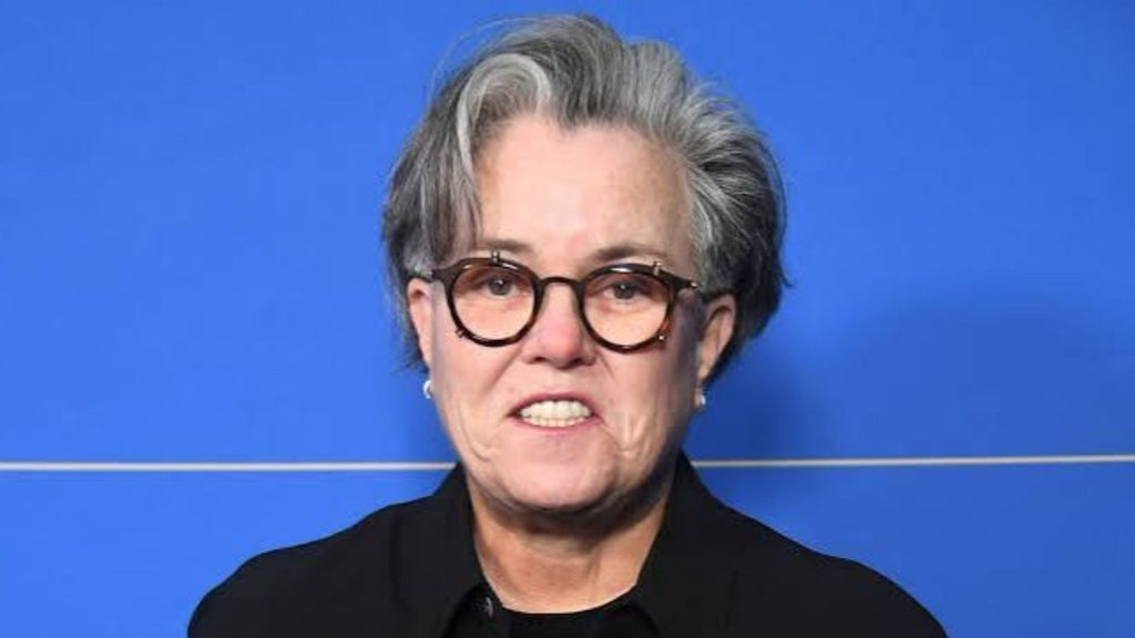 Rosie O'Donnell smiling with short grey hair, round glasses, and a black shirt against blue backdrop.