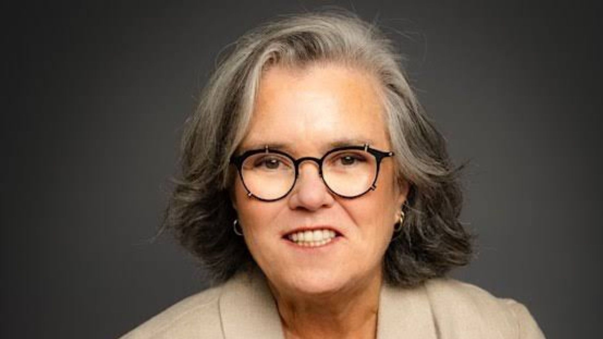 Headshot of Rosie O'Donnell with short gray hair and clip-on flip-up glasses, smiling while wearing a beige blazer against a dark grey background.