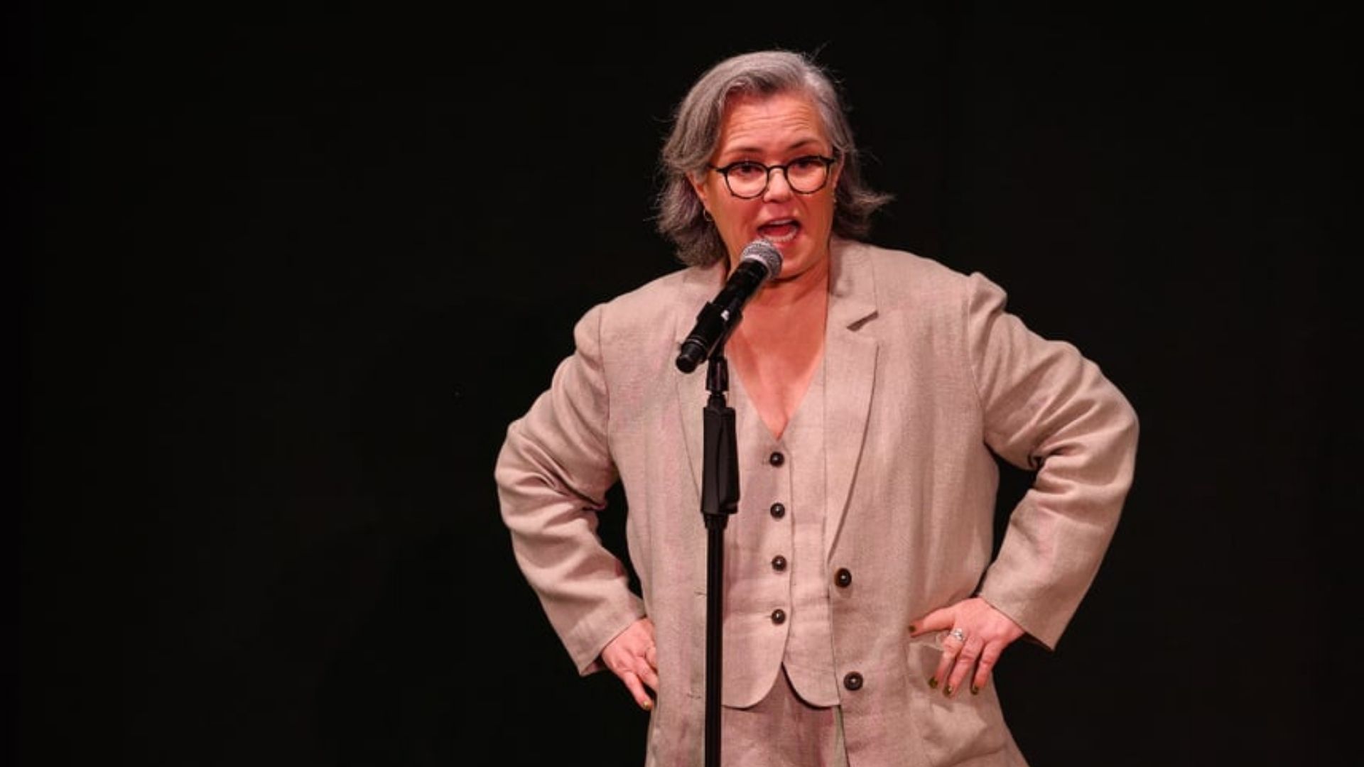 Comedian Rosie O'Donnell speaking into a microphone on stage, wearing a beige three-piece suit, black-rimmed glasses, and standing with hands on her hips against a dark background.
