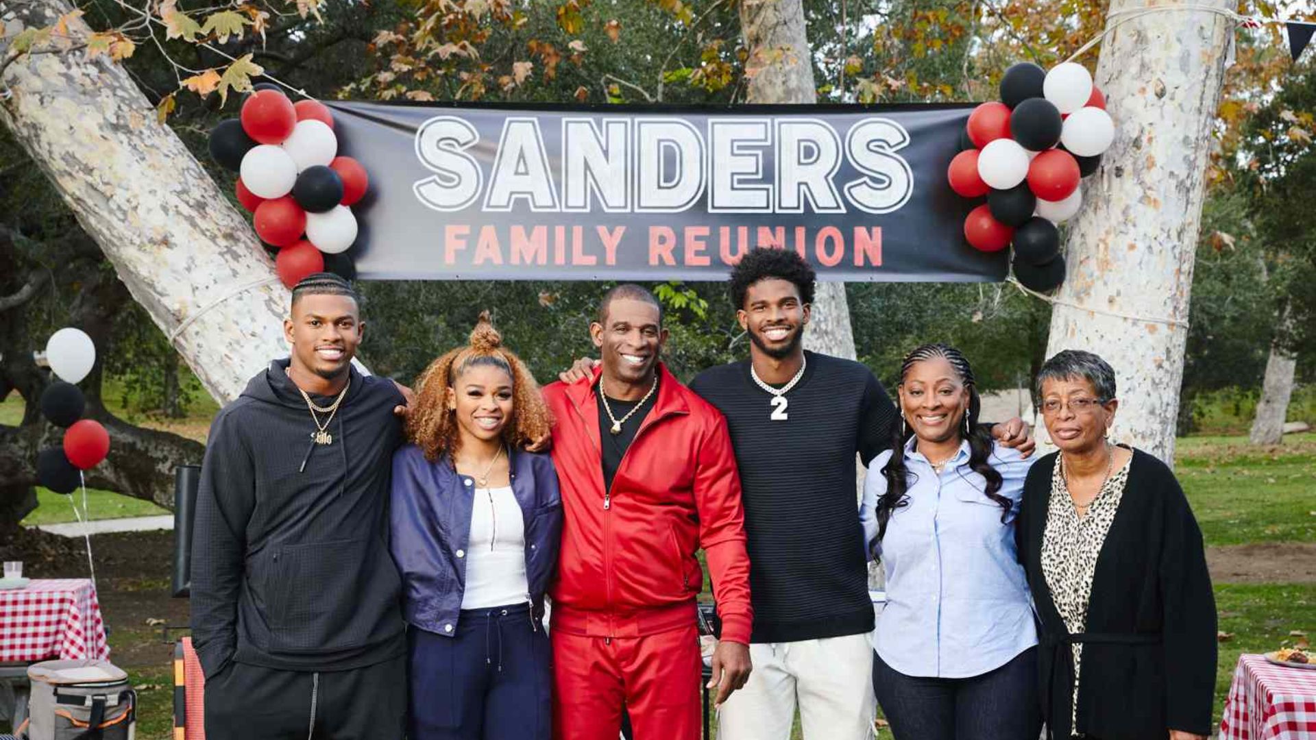 Deion Sanders posing with family members beneath a Sanders Family Reunion banner at an outdoor gathering with balloons and trees.