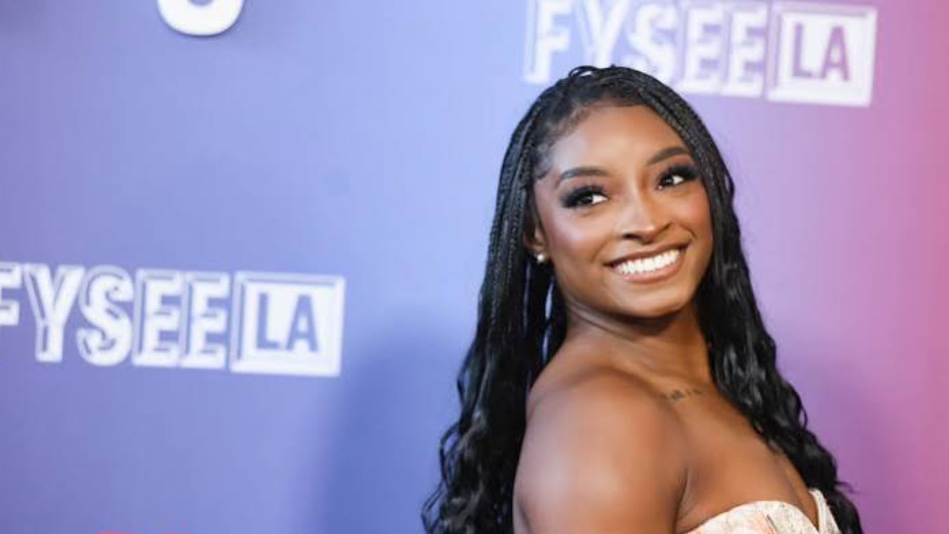 Simone Biles smiling with braided hair in front of a blue and purple Netflix FYSEE LA backdrop.
