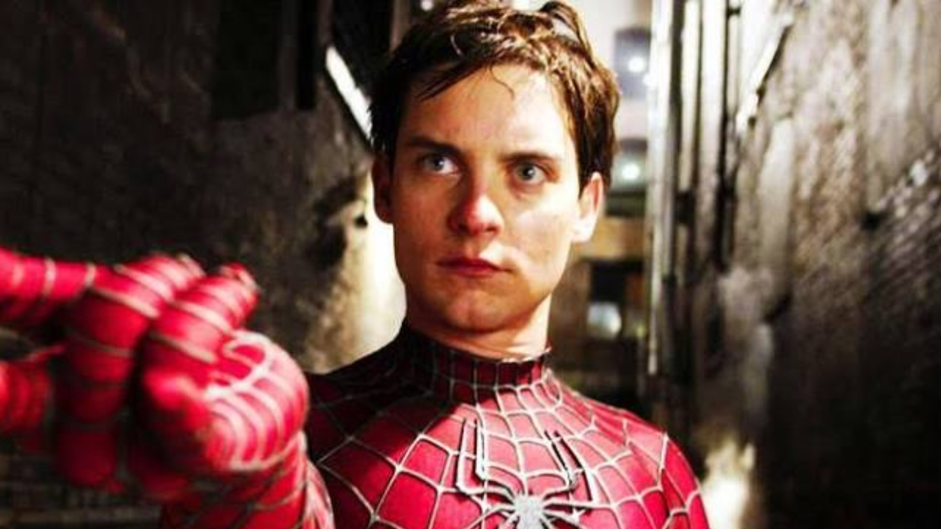 tobey maguire in red spider suit with web pattern and raised gloved hand standing in a narrow dimly lit urban alleyway at night