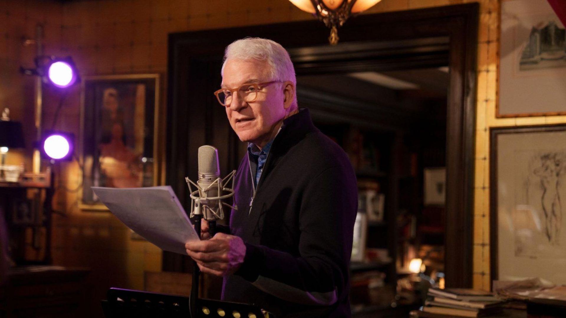 Steve Martin reading from a script while recording at a microphone in a warmly lit studio.