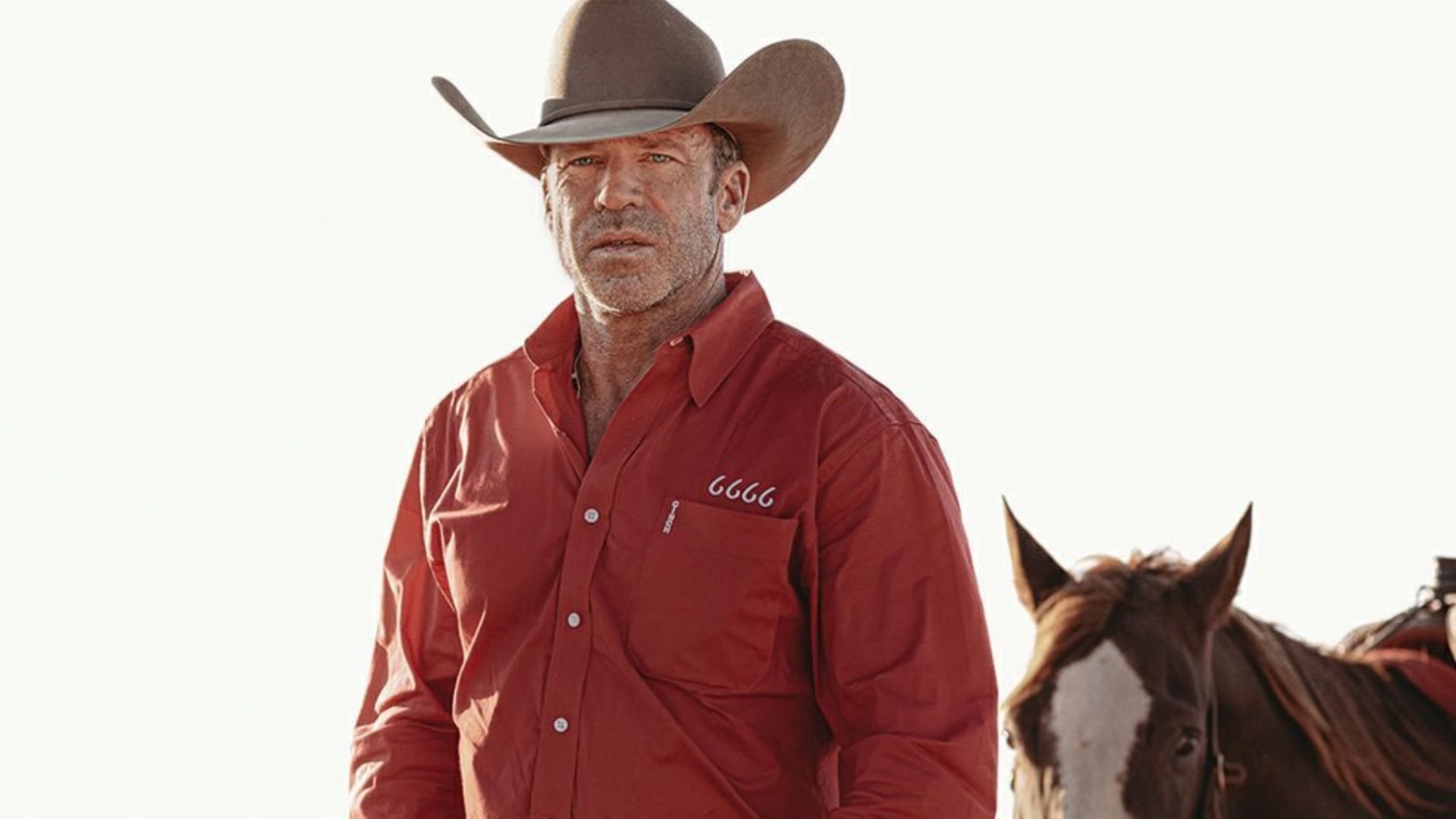Taylor Sheridan wearing a brown cowboy hat and red shirt while standing beside a saddled horse outdoors.