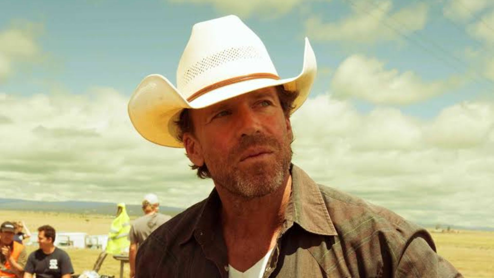 Taylor Sheridan wearing a white cowboy hat outdoors in an open field under a cloudy blue sky.