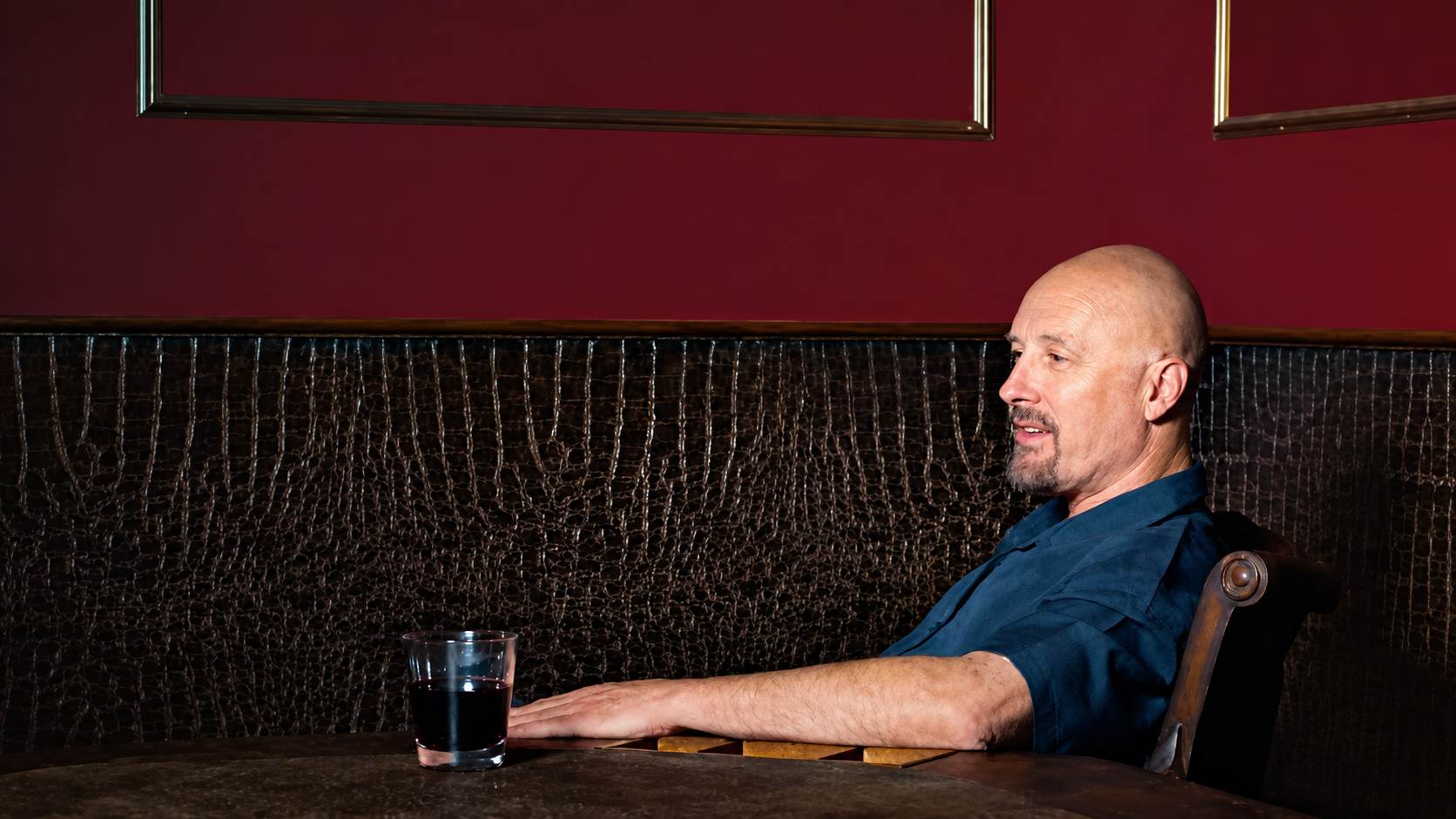 Jim Jannard sitting in a dark lounge booth wearing a blue shirt with a glass on the table and warm indoor lighting