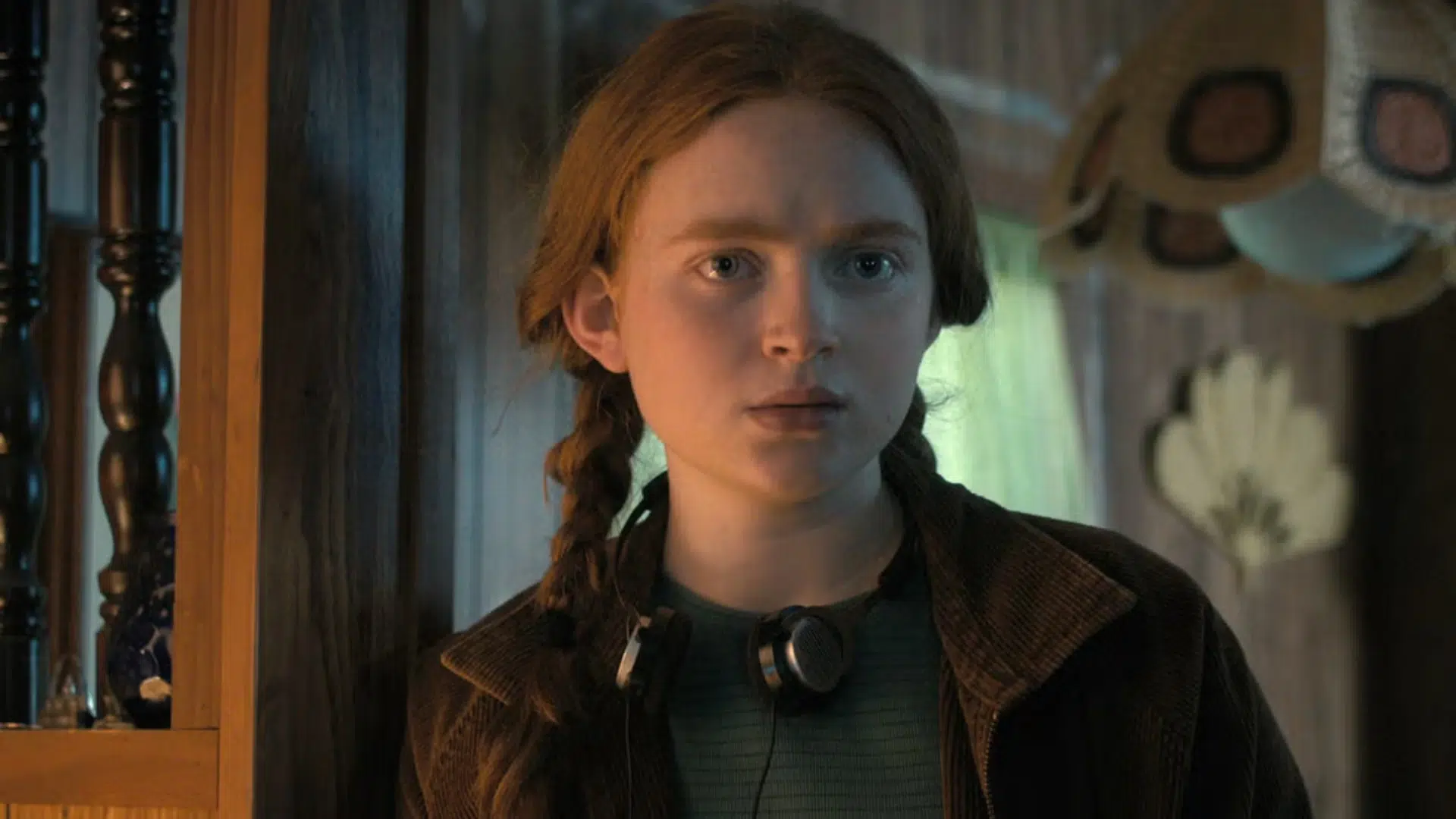 young girl with braided hair wearing a brown jacket and green shirt in a dimly lit room with hats on the wall