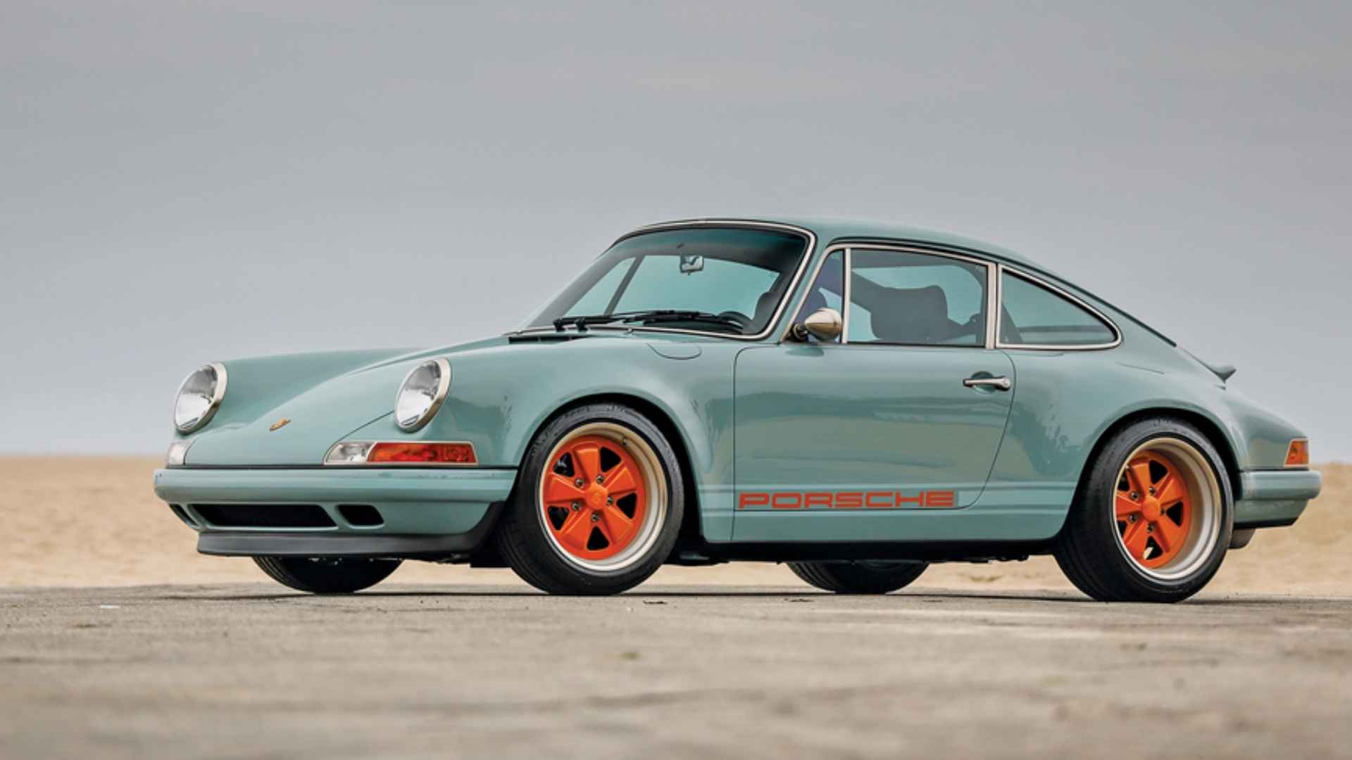 Pale blue Porsche 911 reimagined by Singer with orange wheels and side lettering parked on pavement.