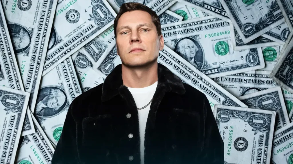 man in a black jacket standing in front of a backdrop covered with overlapping us dollar bills