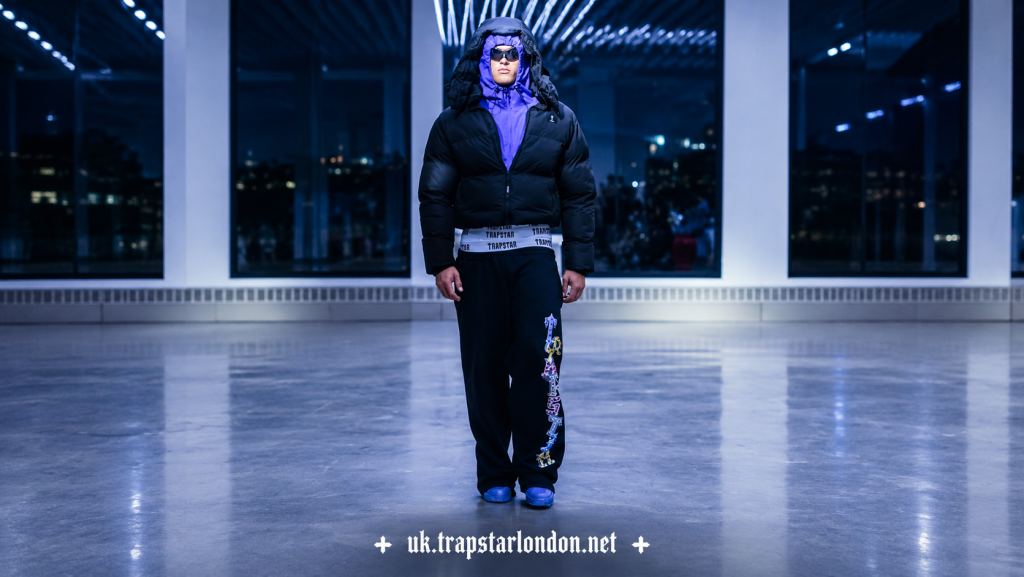 Person in black puffer jacket with hood indoors at night with large windows in background