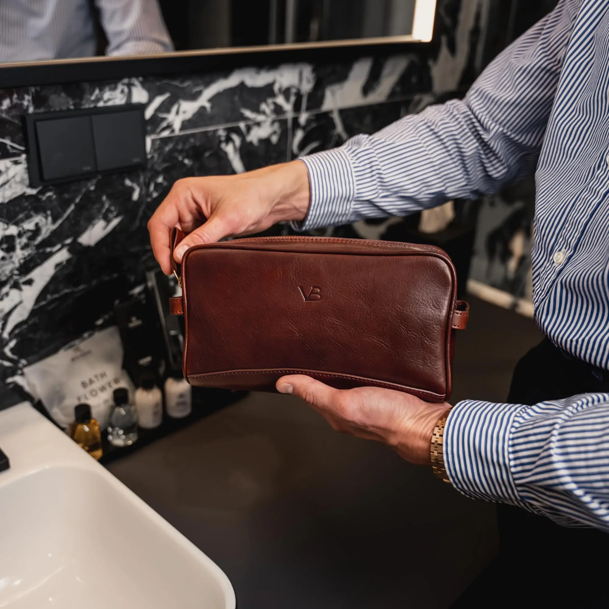 Brown leather toiletry bag held by person in striped shirt in bathroom setting