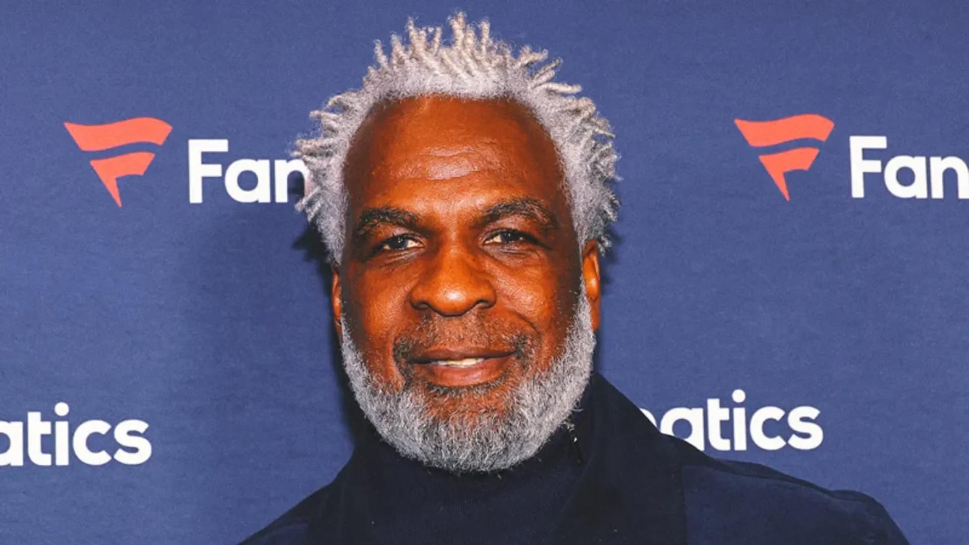 charles oakley with white hair and beard posing at an event with a blue Fanatics-branded backdrop behind him